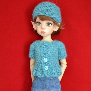 Könnte beinhalten: Ein blauer Strickcardigan mit Knöpfen, getragen von einer Puppe mit einer blauen Strickmütze und einem blauen Jeansrock.