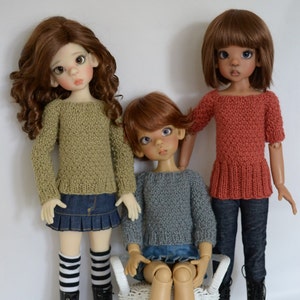 Puede incluir: Tres muñecas con suéteres de punto. La muñeca de la izquierda lleva un suéter verde y una falda azul. La muñeca del medio lleva un suéter gris y una falda azul. La muñeca de la derecha lleva un suéter rojo y pantalones vaqueros azules.