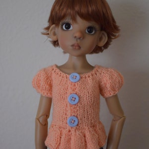 May include: A doll with brown hair and brown eyes wearing a peach knitted sweater with blue buttons and a denim skirt.