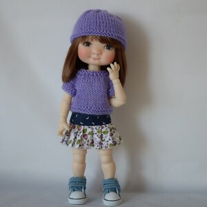 May include: A purple knitted hat and sweater set for a doll. The doll is wearing a white and floral print skirt and blue sneakers.