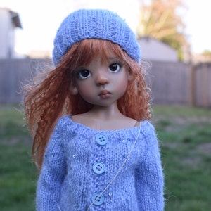 Puede incluir: Una muñeca con el pelo largo, rizado, de color marrón rojizo que lleva un gorro de punto azul claro y un cárdigan de punto azul claro con botones plateados y un collar de cadena plateada.
