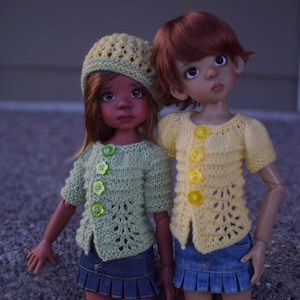 Puede incluir: Dos muñecas con cárdigans de punto y faldas vaqueras. Una muñeca tiene la piel morena y lleva un gorro de ganchillo amarillo. La otra muñeca tiene la piel clara y el pelo rojo.