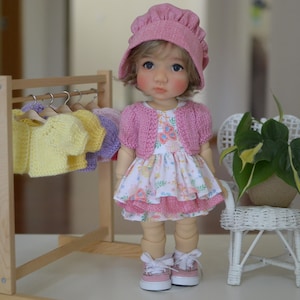 Puede incluir: Una muñeca con gorro de punto rosa, cárdigan a juego y un vestido con estampado floral y ribete rosa. La muñeca lleva zapatillas rosas y está de pie junto a un perchero de madera con suéteres en miniatura amarillos, morados y rosas.
