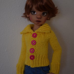 May include: A doll with brown hair wearing a yellow knitted cardigan with pink buttons and a blue denim skirt.