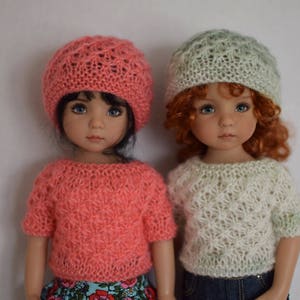 May include: Two dolls wearing knitted hats and sweaters. The doll on the left is wearing a pink sweater and a pink hat. The doll on the right is wearing a white sweater and a green hat.