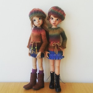 Puede incluir: Dos muñecas con suéteres y sombreros de punto de colores. Las muñecas llevan faldas de mezclilla y botas marrones. Las muñecas están de pie sobre una superficie de madera.