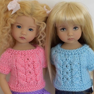 Könnte beinhalten: Zwei Puppen mit blonden Haaren, die Strickpullover tragen. Eine Puppe trägt einen rosa Pullover, die andere einen hellblauen Pullover. Beide Pullover haben ein Zopfmuster. Die Puppen schauen in die Kamera.