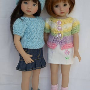 以下が含まれることがあります： 手作りの服を着た2体の人形。1体は青いニット帽とセーター、デニムスカートとスニーカーを着用。もう1体はパステルカラーのセーター、花の付いた白いスカート、白い靴を履いています。