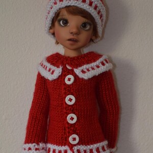 Könnte beinhalten: Ein roter Strickpullover mit weißen Verzierungen und weißen Knöpfen. Der Pullover hat einen weißen Kragen und wird von einer Puppe mit braunen Haaren und einer roten Strickmütze mit weißen Verzierungen getragen.
