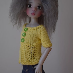 Puede incluir: Una muñeca con pelo gris que lleva un suéter de punto amarillo con botones verdes y una falda vaquera azul.