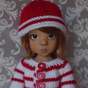 Puede incluir: Una muñeca con un suéter de punto a rayas rojas y blancas con botones rojos y un gorro de punto rojo y blanco.