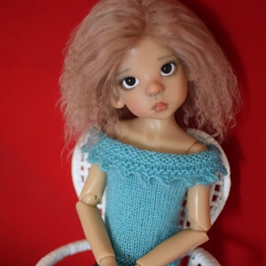 May include: A doll with long, wavy, light pink hair wearing a blue knitted off-the-shoulder top and blue jeans. The doll is sitting on a white wicker chair against a red background.