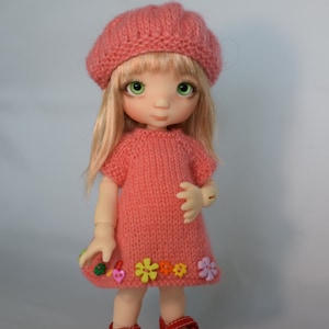 May include: A pink knitted doll with blonde hair, wearing a pink knitted dress with colorful flower buttons and red shoes.