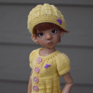 Könnte beinhalten: Eine Puppe mit einem gelben Strickmütze und Pullover mit rosa Knöpfen und einem lila Blumenakzent. Die Puppe hat braunes Haar und braune Augen.