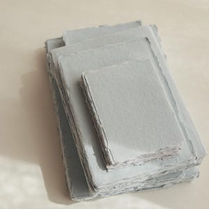 Puede incluir: Una pila de hojas de papel artesanal azul claro con bordes festoneados. El papel es texturizado y tiene una superficie ligeramente rugosa.