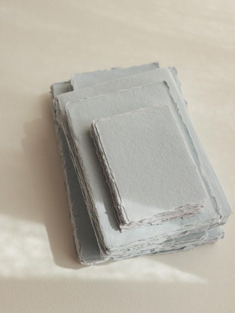 Puede incluir: Una pila de hojas de papel artesanal azul claro con bordes festoneados. El papel es texturizado y tiene una superficie ligeramente rugosa.