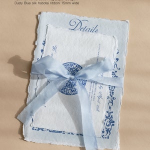 Puede incluir: Una invitaci&oacute;n de boda azul y blanca con una cinta azul atada alrededor. La invitaci&oacute;n est&aacute; impresa en papel texturizado y dice "Detalles" en una fuente cursiva. La invitaci&oacute;n est&aacute; atada con una cinta azul y tiene un c&oacute;digo QR.