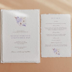May include: A wedding invitation suite with a floral design in shades of purple and white. The invitation card features the text "Please join us to celebrate the wedding of Noah & Charlotte 13 September 2023". The details card includes the text "Details" and lists the wedding ceremony and reception information.