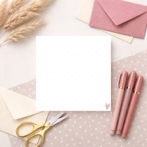 Può includere: Un blocco note quadrato bianco con piccoli pois grigi e un cuore rosa nell'angolo. Tre penne rosa, forbici dorate e due buste sono disposte attorno al blocco note su una superficie a pois. Steli di grano essiccati nell'angolo in alto a sinistra.