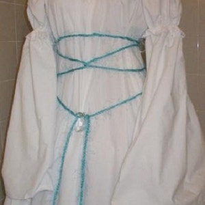 Op de afbeelding: Een witte off-the-shoulder jurk met lange, volumineuze mouwen. De jurk heeft een turquoise kruisontwerp rond de taille en een decoratieve hanger. De mouwen hebben gerimpelde manchetten. De jurk is gemaakt van een zachte stof.