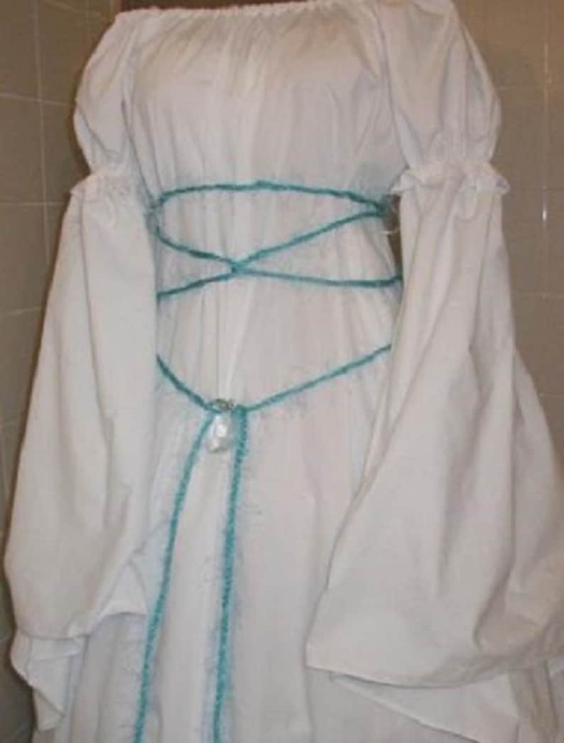 Op de afbeelding: Een witte off-the-shoulder jurk met lange, volumineuze mouwen. De jurk heeft een turquoise kruisontwerp rond de taille en een decoratieve hanger. De mouwen hebben gerimpelde manchetten. De jurk is gemaakt van een zachte stof.