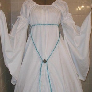Op de afbeelding: Een witte jurk in middeleeuwse stijl met lange, vloeiende mouwen. De jurk heeft een ronde hals en een turquoise geborduurde riem die de taille accentueert. Een decoratieve sluiting bevindt zich in het midden van de turquoise riem.