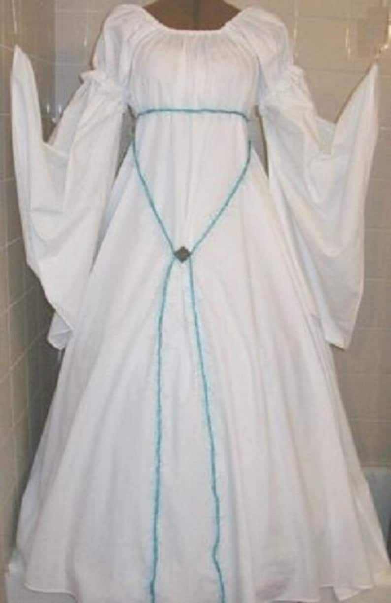 Op de afbeelding: Een witte jurk in middeleeuwse stijl met lange, vloeiende mouwen en een ronde halslijn. De jurk heeft een turquoise rand rond de taille en de voorkant, met een decoratieve sluiting in het midden. De jurk is gemaakt van een zachte, vloeiende stof.