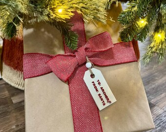 Könnte beinhalten: Ein Geschenk unter einem Weihnachtsbaum. Das Geschenk ist in braunes Papier gewickelt und mit einem rot-weiß karierten Band gebunden. Ein Anhänger lautet: "To: Archers From: Santa". Der Baum hat Lichter.