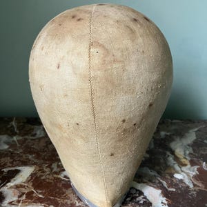 May include: A vintage wig or hat form, featuring a rounded head shape and a tapered neck. The form is covered in a light tan fabric with dark spots and a silver metal base. The form is likely used for displaying or shaping hats and wigs.