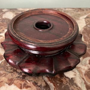 May include: A dark red, wooden pedestal with a circular top and a flared, petal-like base. The top has a central hole. The wood grain is visible, and the finish is glossy. This decorative stand is suitable for displaying small objects.