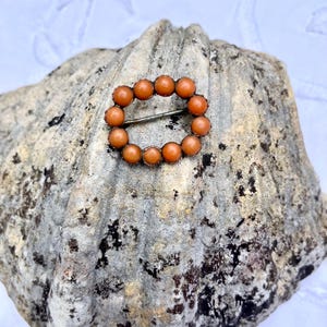 May include: A vintage brooch with an oval design, composed of small, orange beads set in a silver-toned metal frame. The brooch is displayed on a textured, grey surface. The beads are approximately 0.5 cm in diameter.