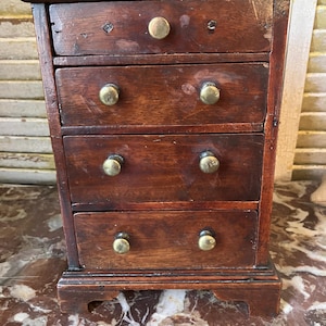 May include: A small, antique wooden chest of drawers with four drawers. The chest is made of dark brown wood and features brass knobs. The piece has a classic, vintage aesthetic, and is likely used for storing small items.