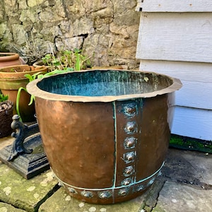 May include: A large, hammered copper planter with a decorative band of metal studs around the top and bottom. The planter has a greenish patina and is sitting on a stone patio.