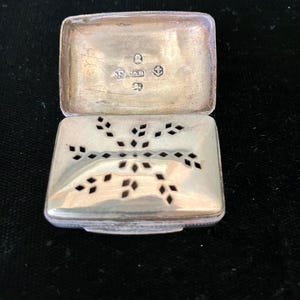 May include: Open silver rectangular box with a hinged lid. The lid is open, revealing the interior and hallmarks. The top of the box has a decorative cut-out design. The box is on a black surface.