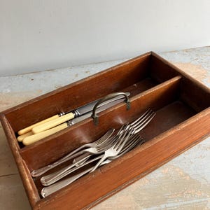 Könnte beinhalten: Eine hölzerne Besteckschublade mit Metallgriff, die Besteck enthält. Die Schublade ist in Fächer unterteilt, die Gabeln und Messer mit cremefarbenen Griffen aufnehmen. Das Holz hat einen warmen, rotbraunen Farbton.