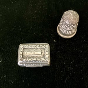 May include: A silver thimble and a silver rectangular box with an engraved design. The box has a raised inscription on the top.