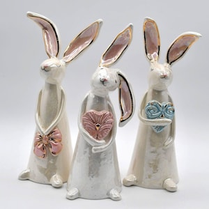 May include: Three white ceramic bunny figurines with pink and gold accents. Each bunny is holding a heart-shaped object. One bunny is holding a pink heart, one is holding a blue heart, and one is holding a pink heart with gold accents.