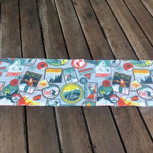 May include: A white fabric table runner with a colorful pattern of vintage ski resort logos. The logos feature names like Zermatt, Norge, Vail, and Lake Tahoe. The runner is perfect for adding a touch of wintery charm to your table decor.