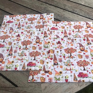 May include: Set of three white quilted placemats with a fall theme. The placemats feature a repeating pattern of gnomes, trees, pumpkins, and sunflowers. The placemats are trimmed with a red and white checkered border.
