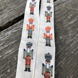May include: Two white ribbons with a repeating pattern of nutcracker figures. The nutcrackers are wearing red, gold, and black uniforms with white accents. The ribbons are on a wooden surface.
