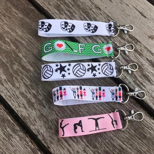 May include: Five different keychains with colorful ribbon straps and silver key rings. The ribbons feature various designs, including a heart, stars, a volleyball, and a nutcracker.