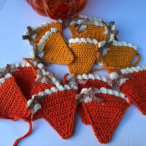 May include: A collection of triangular crochet ornaments shaped like pie slices in shades of orange and gold. Each piece features a white crocheted border along the top edge and is decorated with a small brown and white checkered fabric bow.