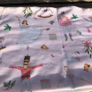 May include: Pink fabric with a repeating pattern of a ballerina, a nutcracker, a rocking horse, a swan, a mouse, and a sprig of mistletoe. The fabric is decorated with a watercolor-like design.