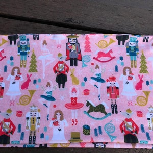 May include: Pink fabric with a repeating pattern of nutcrackers, ballerinas, rocking horses, and Christmas trees. The fabric has a white background with a pink and green pattern.