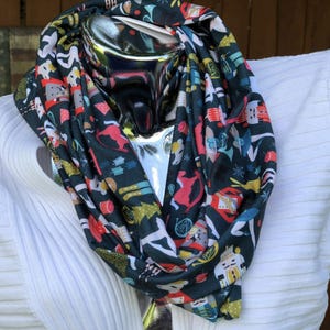 May include: A dark green infinity scarf with a colorful pattern of people, trees, and snowflakes. The scarf is made of a soft, comfortable fabric and is perfect for keeping warm in the winter.