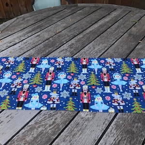 May include: A blue table runner with a repeating pattern of nutcrackers, ballerinas, candy canes, snowflakes, and Christmas trees. The pattern is in shades of red, green, white, and gold.