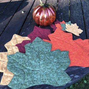 May include: A collection of leaf-shaped placemats in various fall colors, including green, orange, burgundy, and tan. A decorative, shiny, orange pumpkin sits atop a wooden table, complementing the autumn theme. The placemats are quilted.