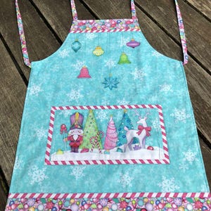May include: A light blue Christmas apron with a festive design. The apron features a snowflake pattern and a rectangular panel with a nutcracker, trees, and a reindeer. Colorful ornaments and a striped border complete the design. The apron has straps for the neck and waist.