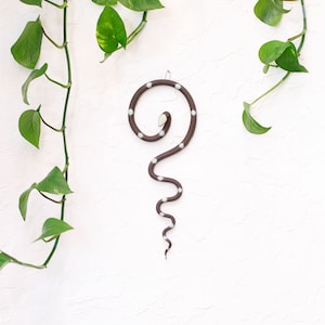 May include: A brown ceramic snake with white polka dots hanging on a white wall. The snake is curled in a coil shape with its tail hanging down.
