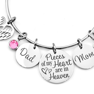 Könnte beinhalten: Ein silbernes Armband mit einem herzförmigen Engelsflügel-Anhänger, zwei Anhängern mit den Worten "Dad" und "Mom" und einem Anhänger mit den Worten "Pieces of my Heart are in Heaven". Das Armband hat rosa und grüne Kristalle.